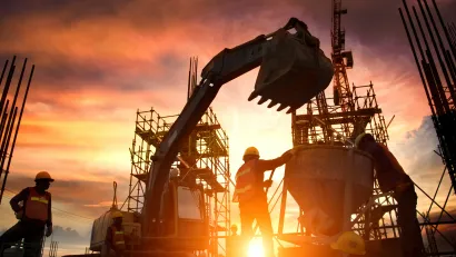 Construction workers, bulldozer, and unfinished bridge or highway sillhouetted against orange sunset sky